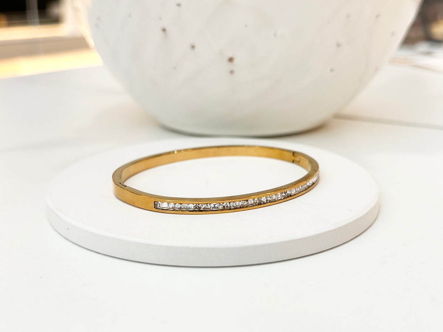 
                  
                    A gold bangle bracelet with a row of small clear stones, displayed on a round white coaster with a white ceramic vase in the blurred background.
                  
                