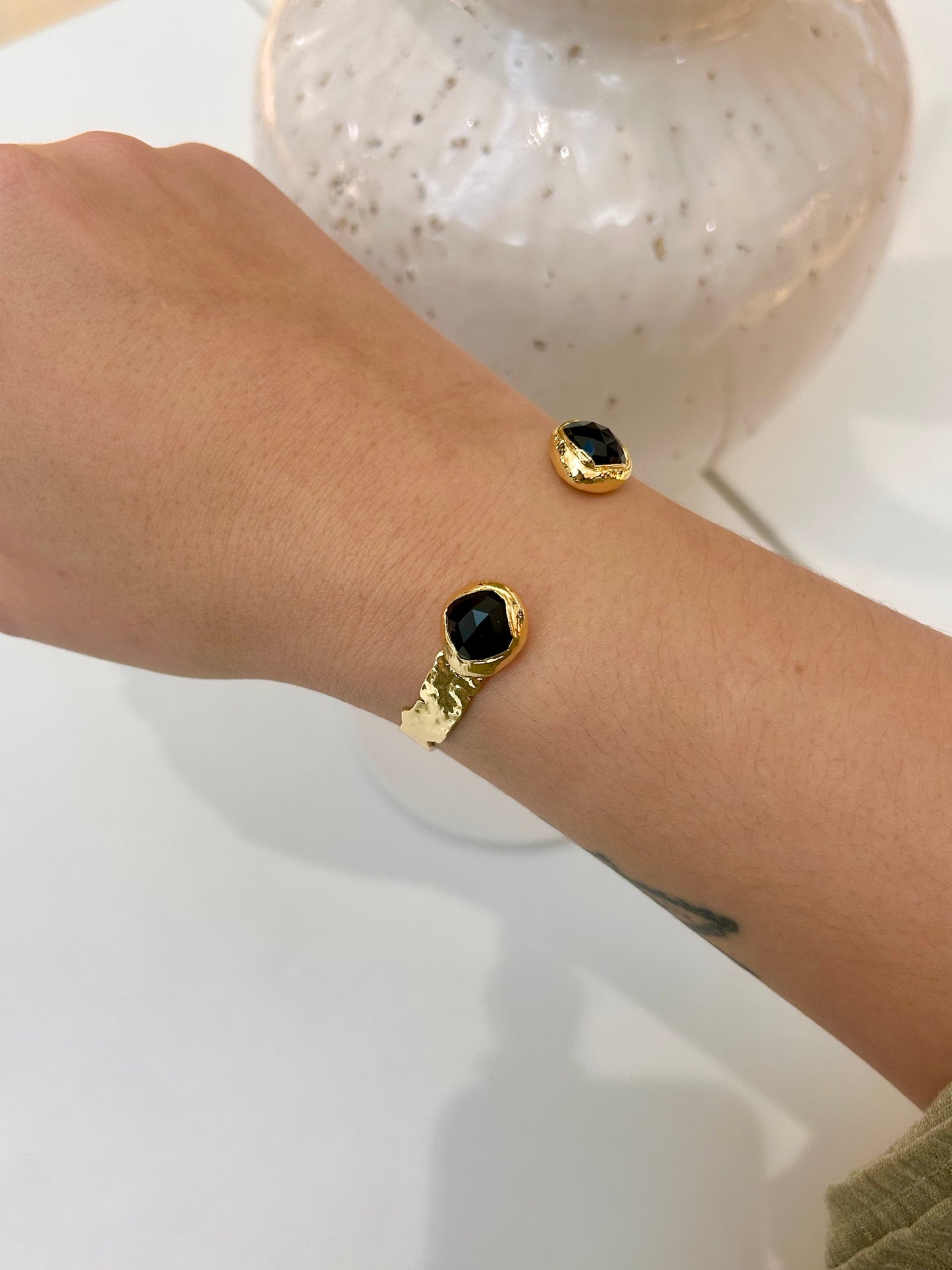 
                  
                    A close-up of a person's wrist wearing two gold-toned bangle bracelets, each with a round black stone, against a light background with a decorative vase.
                  
                