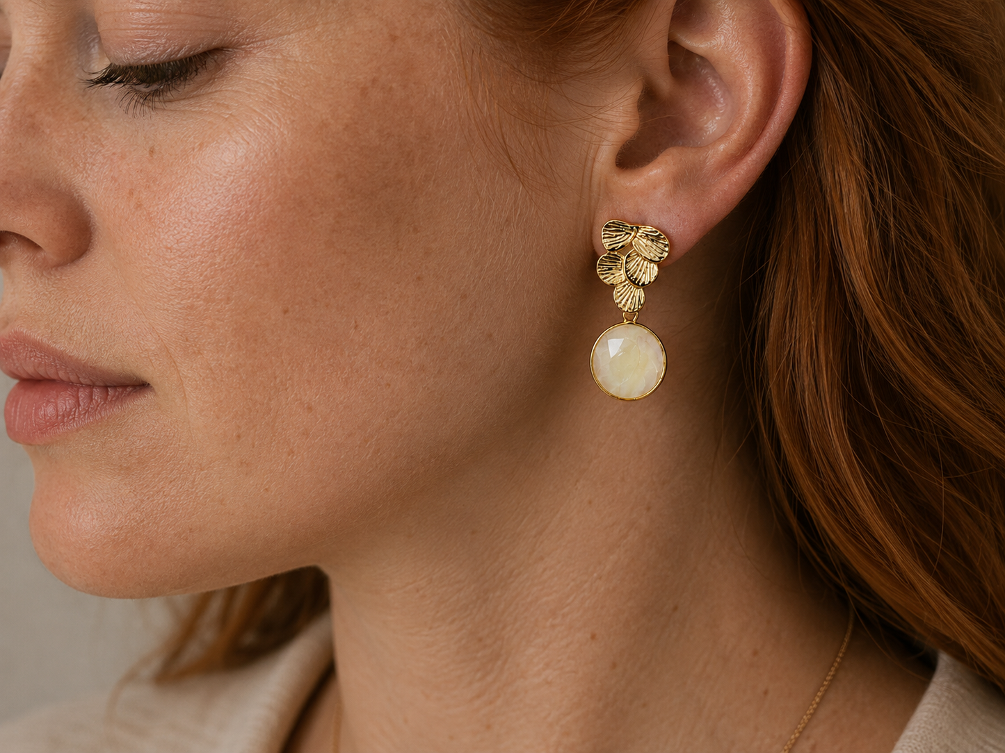 A close-up side profile of a woman wearing a gold dangle earring with a textured leaf cluster and a round cream-colored stone drop.