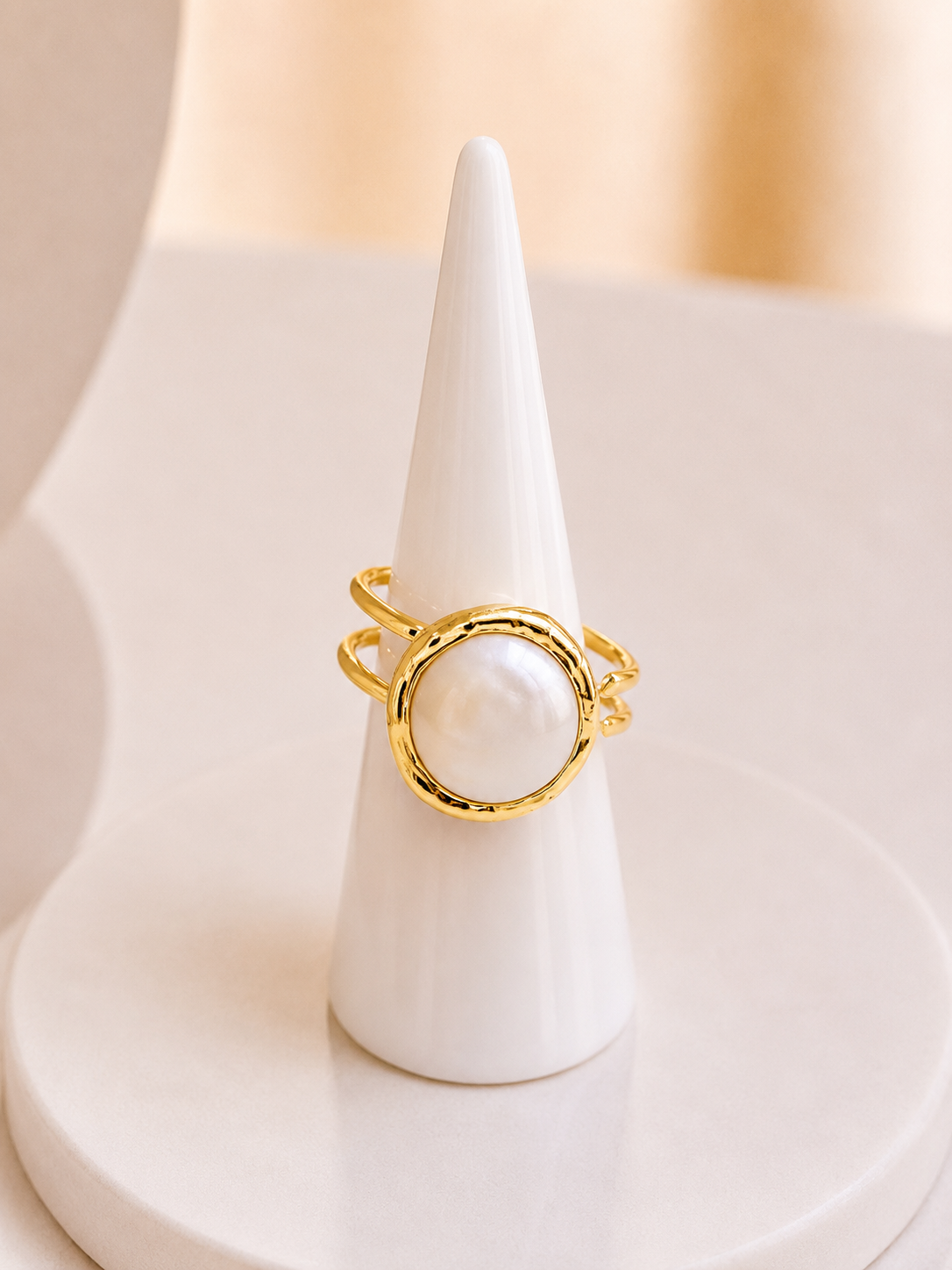 A gold ring with a large white pearl oval center, displayed on a white cone jewellery stand against a soft beige background.