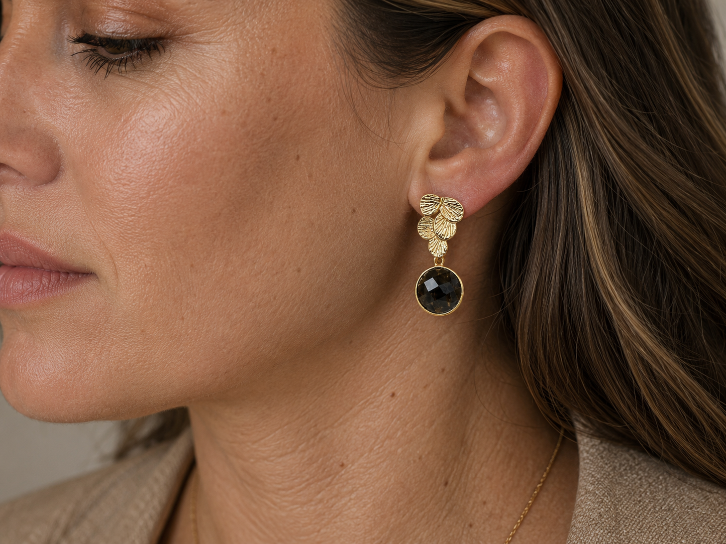 A close-up of a woman's ear wearing a gold dangle earring with a textured gold top and a round dark gray faceted stone.