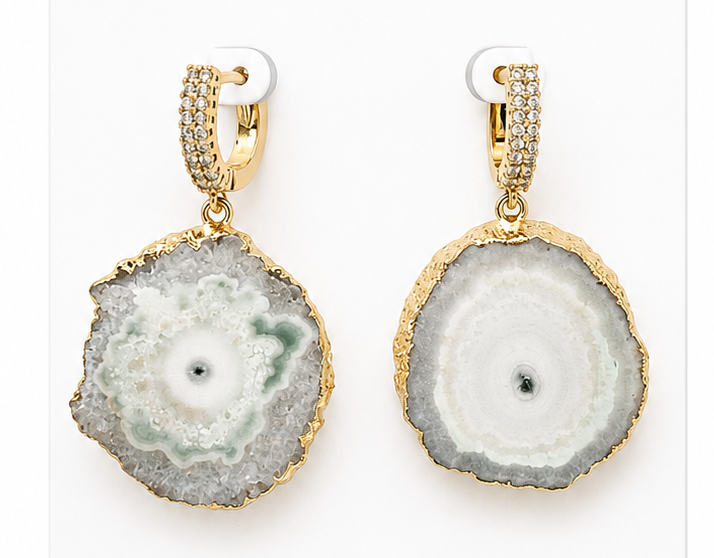 A pair of gold hoop earrings with round agate slice pendants. The left earring features a pale green and white agate pattern, while the right features a white and gray agate pattern, both with gold borders and pavé-set gold hoops.
