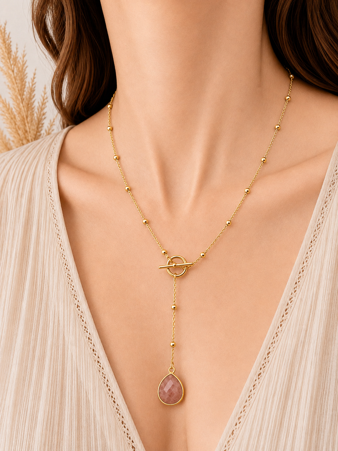 A close-up of a model wearing a delicate gold Y-necklace with a teardrop-shaped sunstone pendant, styled with a beige lace-trimmed top.