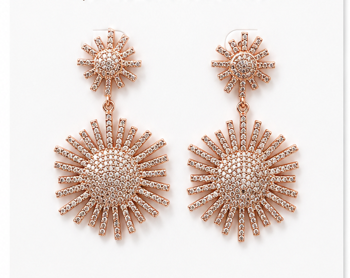 A pair of rose gold dangle earrings with a smaller sunburst stud and a larger sunburst drop, both encrusted with clear rhinestones, shown on a plain white background.