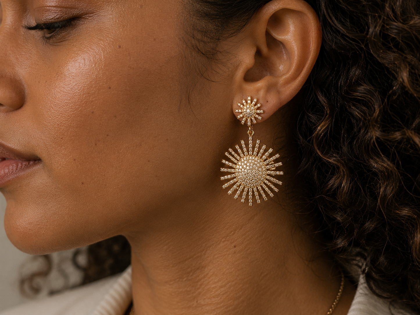 A close-up side profile of a woman wearing a gold-toned drop earring with a sunburst design set with clear stones.