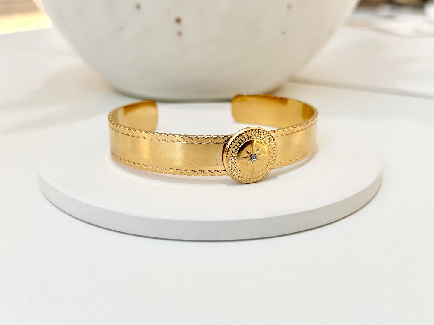 
                  
                    A gold cuff bracelet with textured rope-like detailing and a round decorative clasp, displayed on a white round pedestal.
                  
                