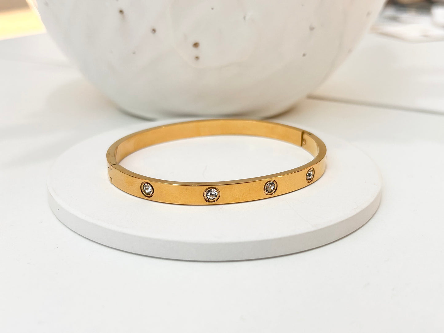 
                  
                    A gold bangle bracelet with five small bezel-set clear stones, displayed on a round white pedestal.
                  
                
