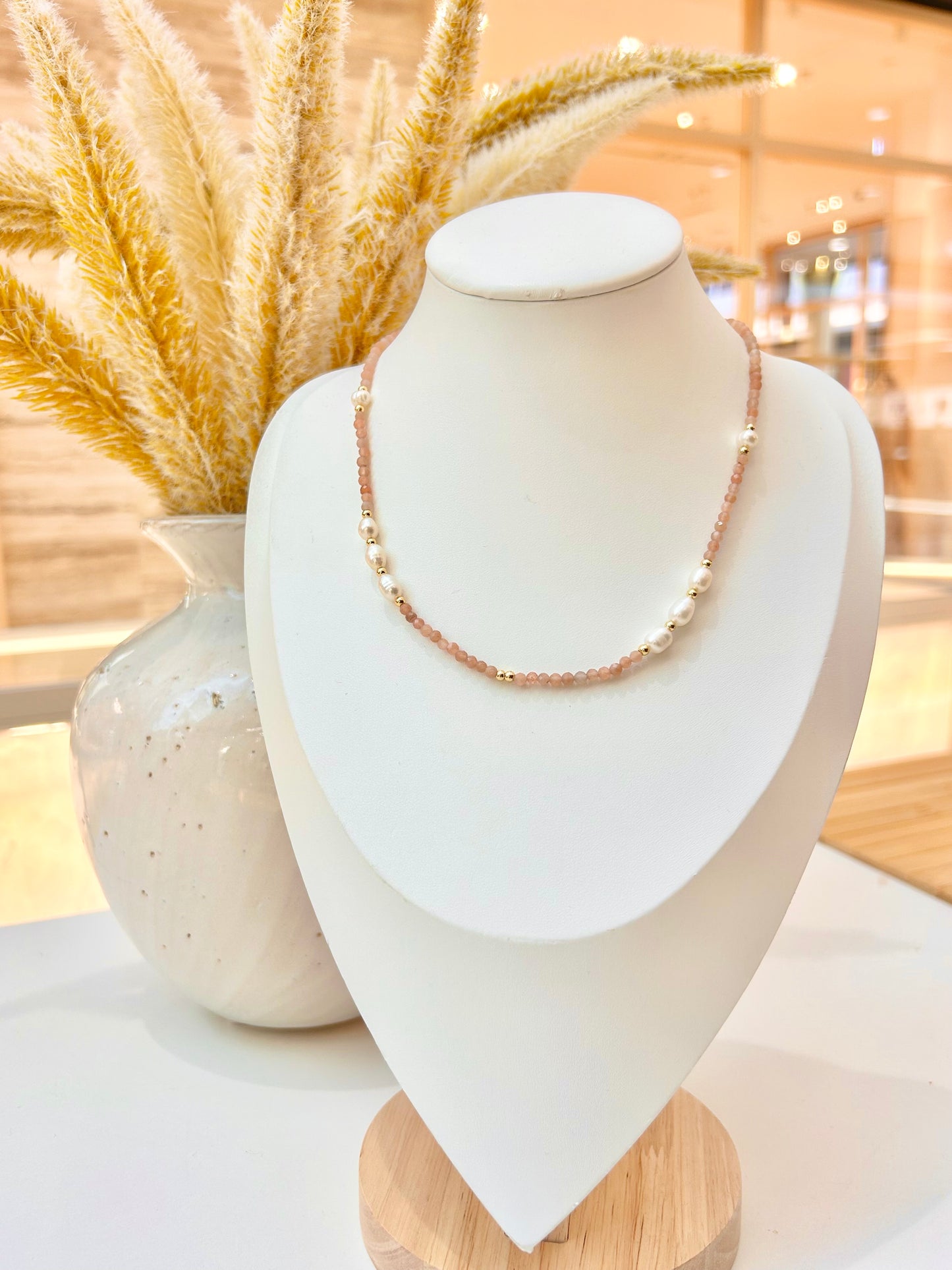 
                  
                    a beaded sunstone necklace with gold accents and freshwater pearls on a white jewellery bust stand
                  
                