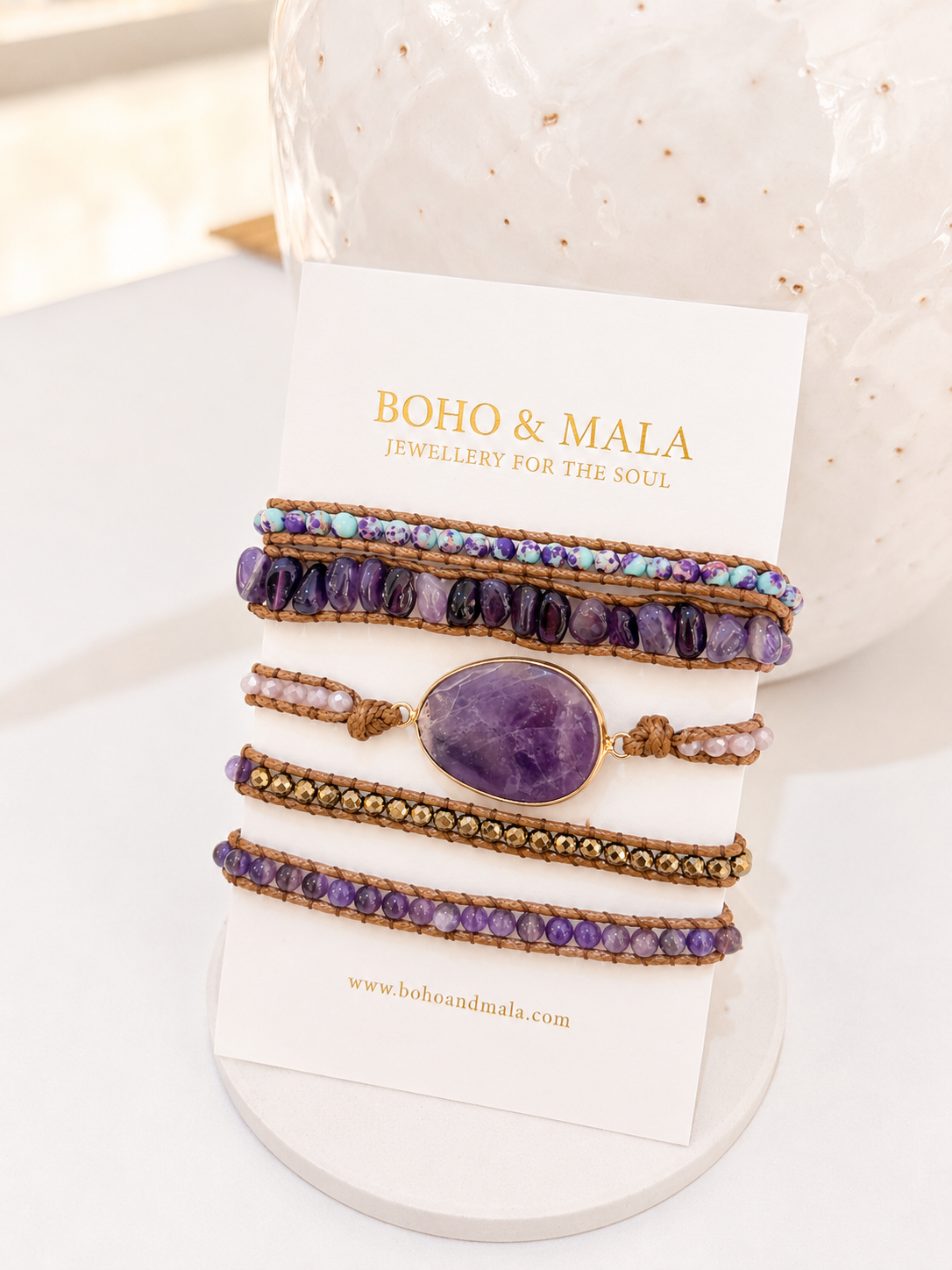 A set of five bohemian-style wrap bracelets from Boho & Mala, featuring purple amethyst beads, a central oval purple stone, and gold-toned metal details, displayed on a white branded card.