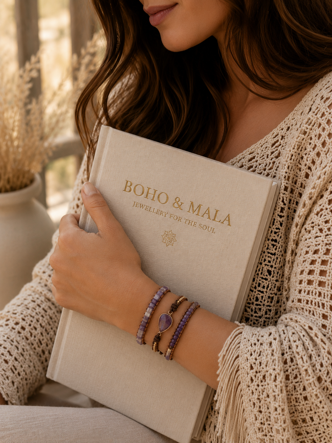 
                  
                    Person holding a book titled 'Boho & Mala' with amethyst wrap bracelet on a blurred background
                  
                