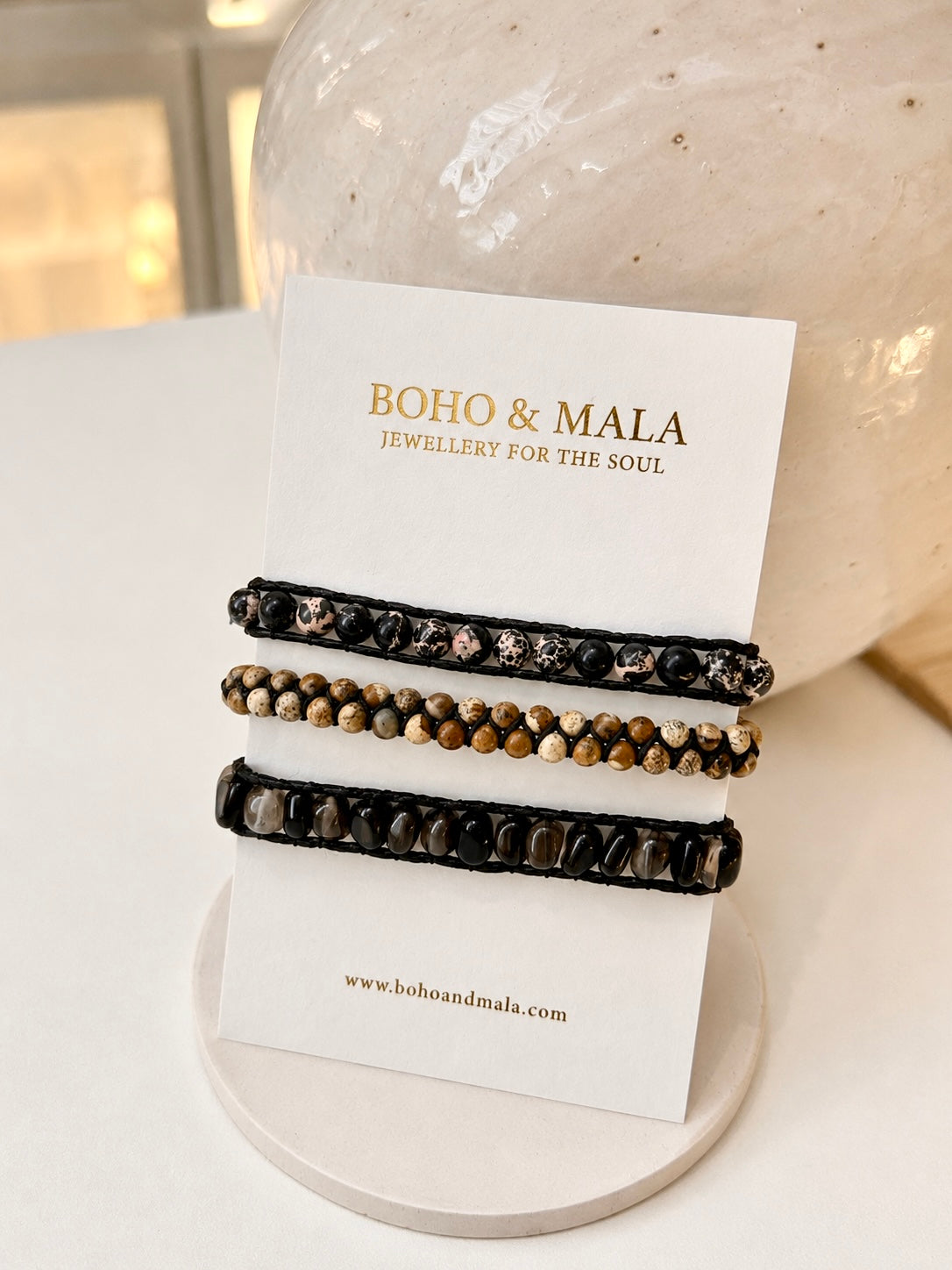 Three beaded natural stone bracelets displayed on a white card stand branded BOHO & MALA. The bracelets feature black cords with black onyx and black agate