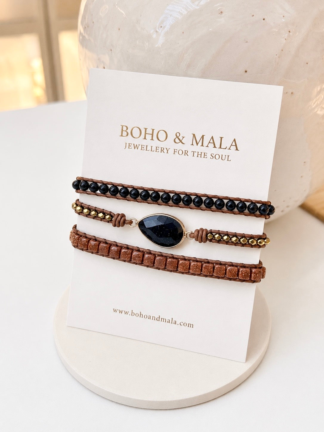 Three bohemian-style bracelets displayed on a white card from Boho & Mala, featuring black onyx mixed beads, brown leather, and a dark teardrop stone, set against a softly blurred background.