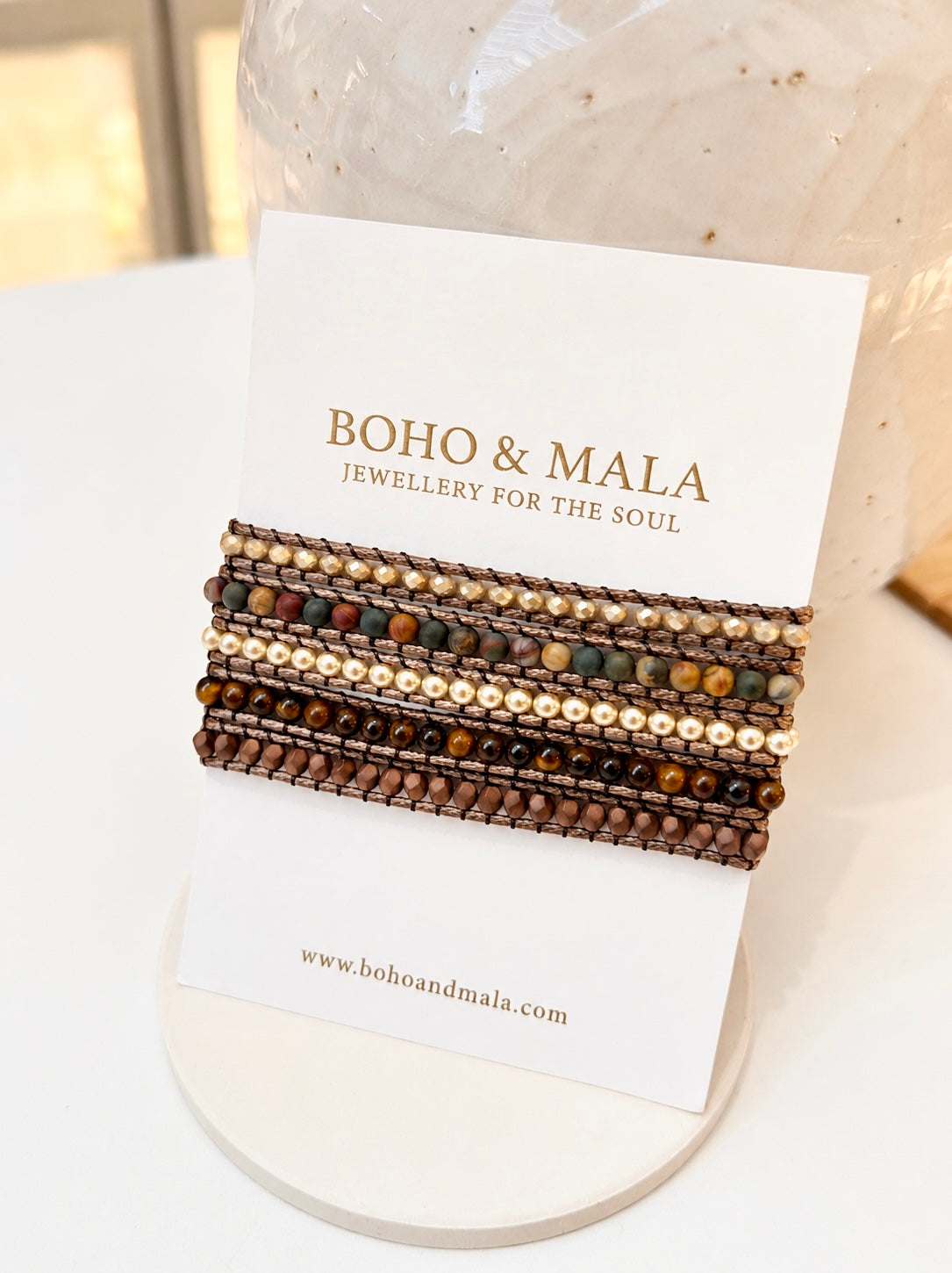 A set of bohemian tigers eye natural beaded bracelets displayed on a white card from the brand BOHO & MALA, resting on a small round white stand.
