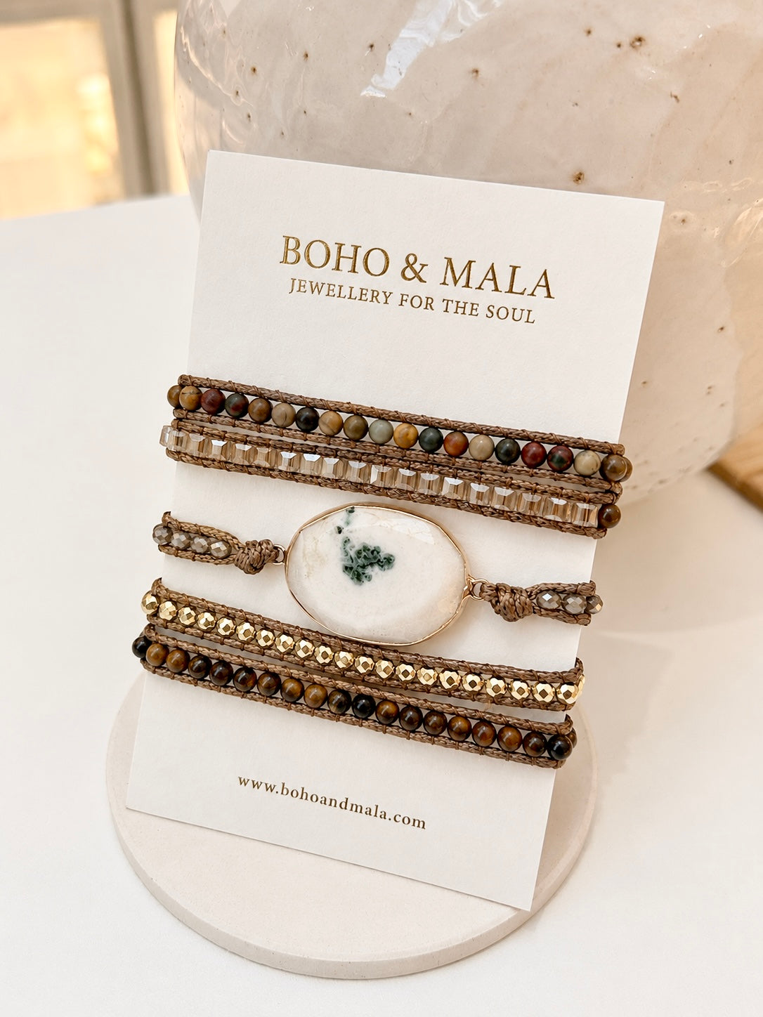 A set of bohemian-style beaded bracelets displayed on a white card stand with the brand name 'BOHO & MALA' printed on it. The bracelets feature brown leather cords, multi-colored beads, clear rhinestones, and a central white agate stone with a green leaf motif.
