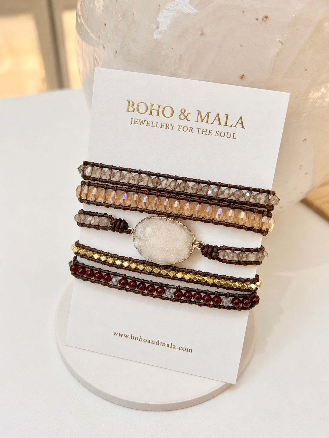 A stack of five bohemian-style beaded bracelets displayed on a white card from the brand BOHO & MALA. The bracelets feature mixed brown, gold, beige, and burgundy natural beads with a central white druzy agate charm, set against a soft-focus background of a white textured vase.