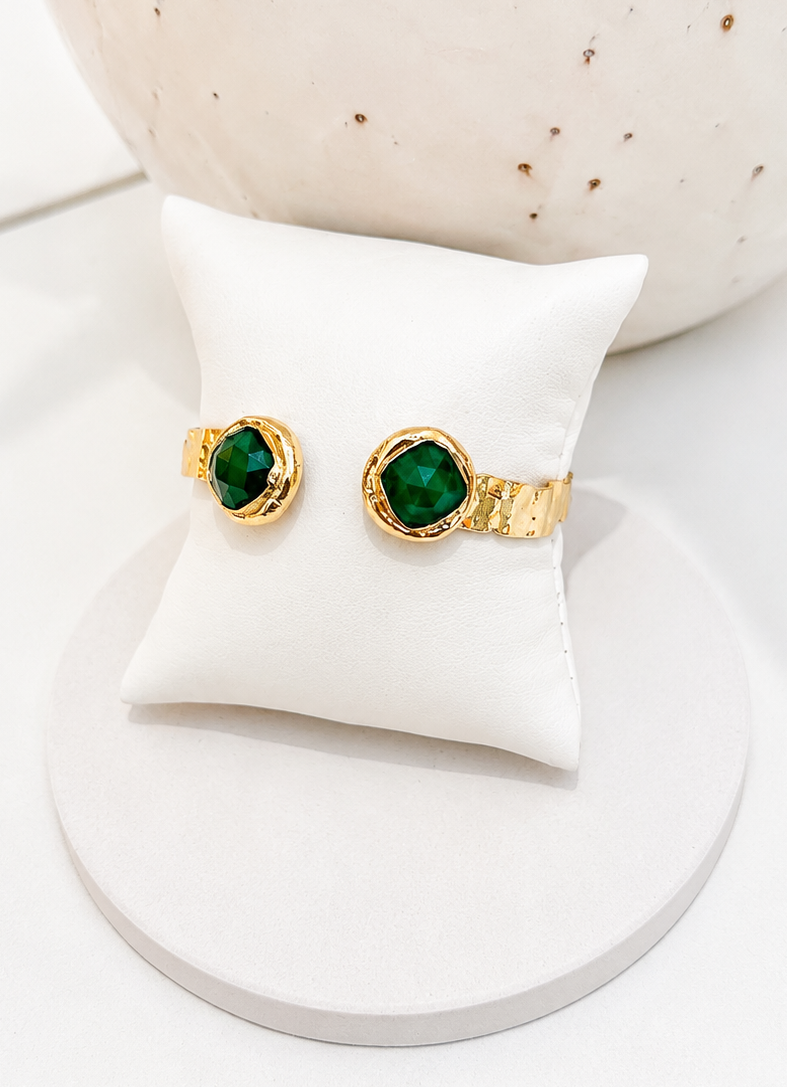 
                  
                    A pair of gold rings with round green gemstones, displayed on a white cushion atop a round white stand.
                  
                