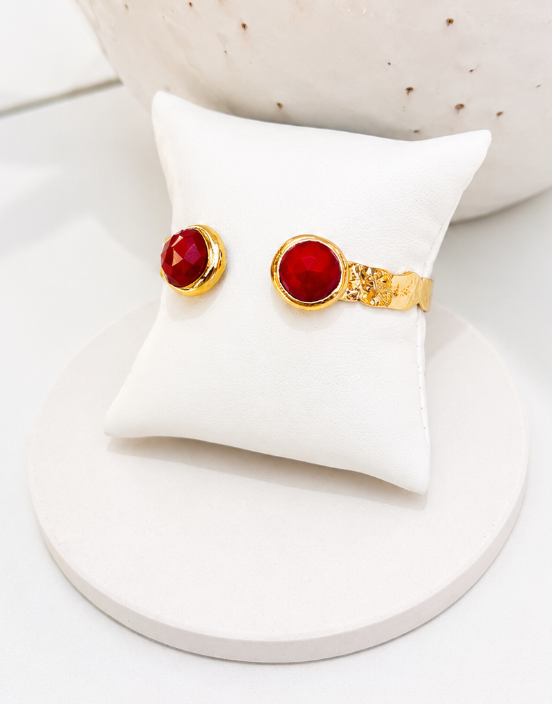 
                  
                    A pair of gold ring-style earrings with round red cabochon stones, displayed on a small white cushion atop a round white stand.
                  
                
