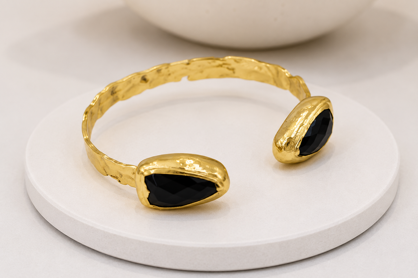 A gold-toned open cuff bracelet with two teardrop-shaped black stone accents, displayed on a round white pedestal.