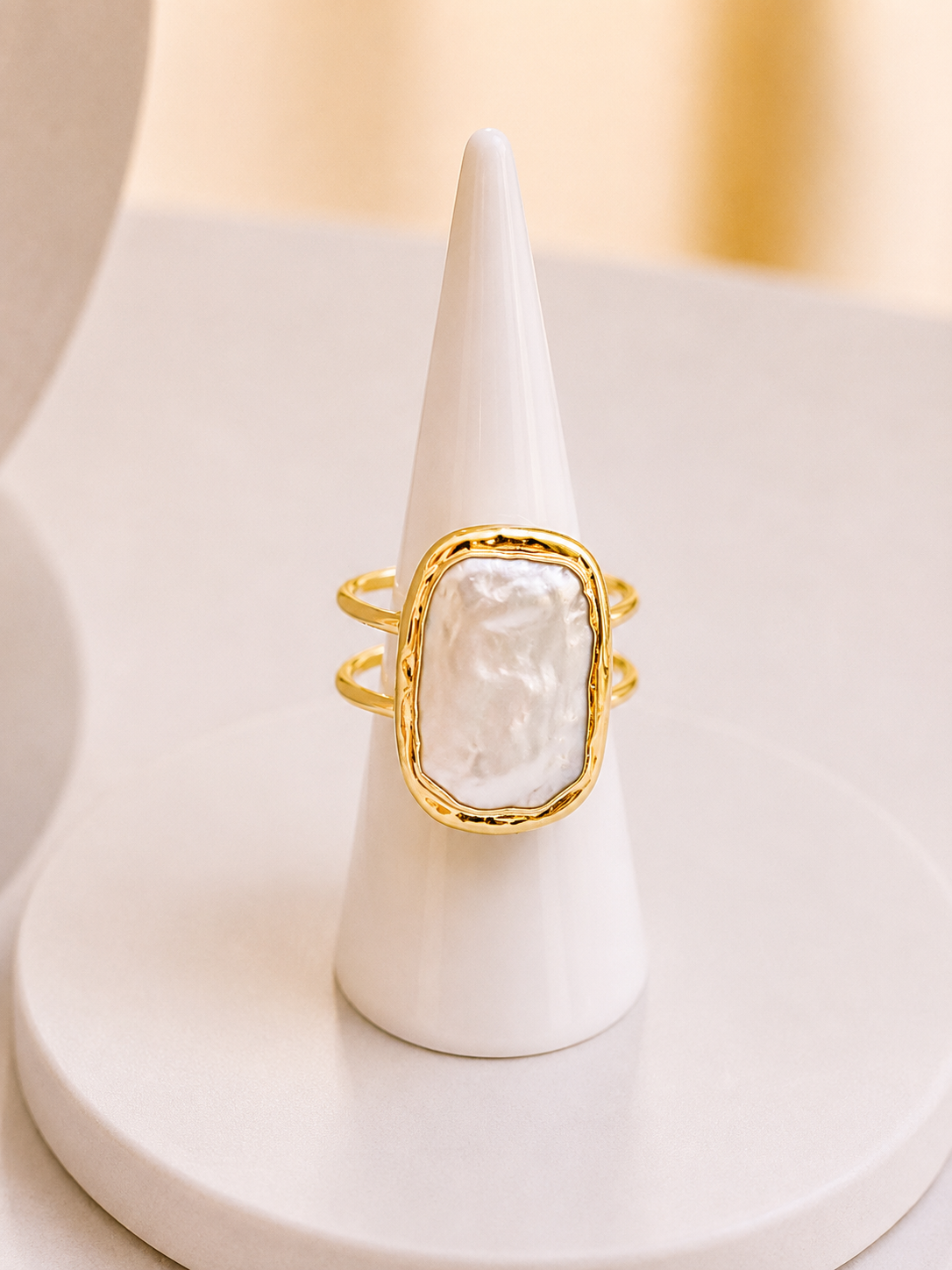 A gold ring with a large, irregular white pearl centerpiece, displayed on a white cone ring stand against a soft beige background.