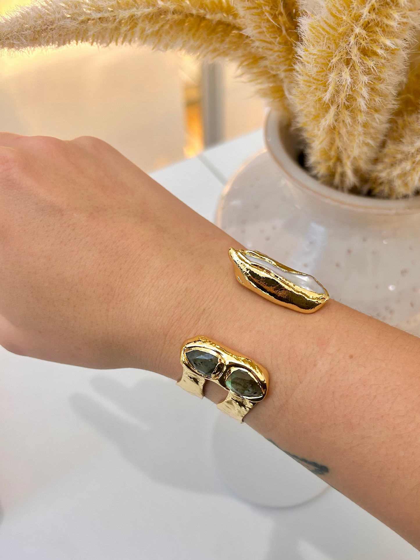 A close-up of a person's wrist wearing two gold-toned cuff bracelets with labradroite stone inlays, set against a bright indoor background with a white vase and dried yellow flowers.