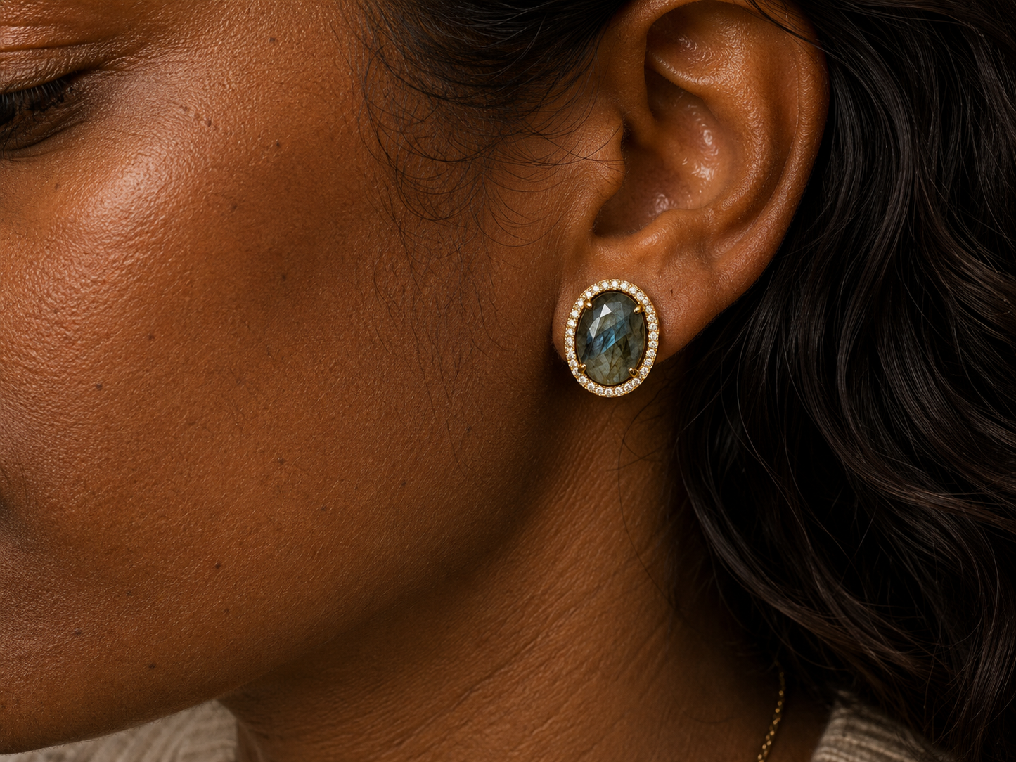Close-up of a person's ear wearing a gold oval stud earring with a dark gray and blue labradorite stone surrounded by a halo of small clear stones.