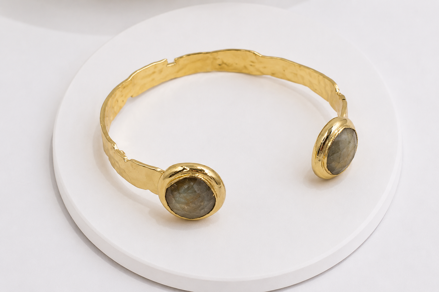 A gold-toned open cuff bracelet with two oval labradorite stones, displayed on a round white pedestal against a white background.