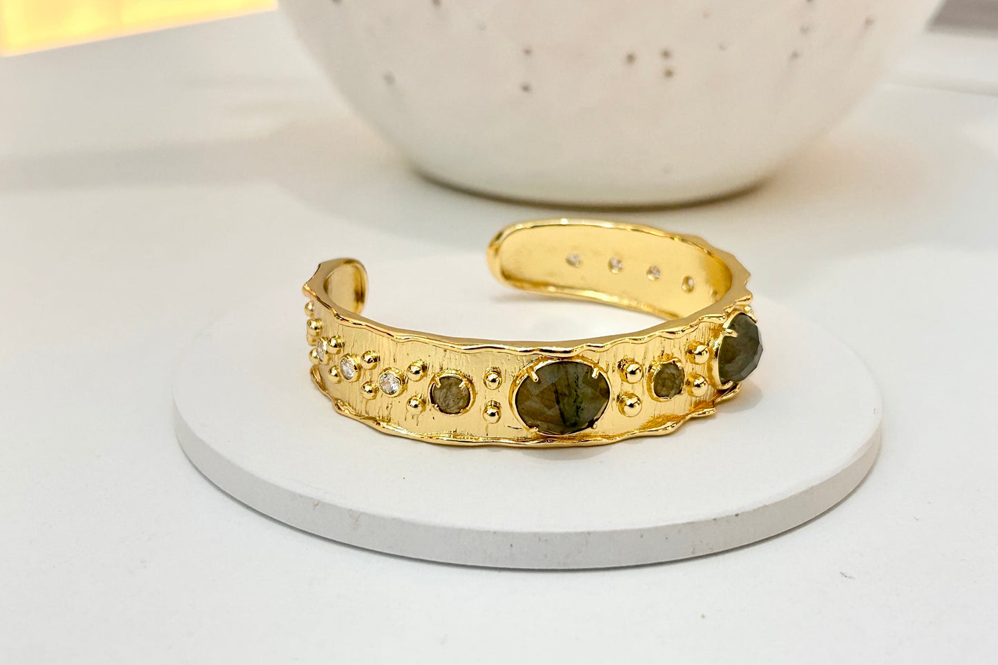 
                  
                    A gold cuff bracelet with textured organic edges and labradorite stones, displayed on a round white coaster with a white speckled vase in the background.
                  
                