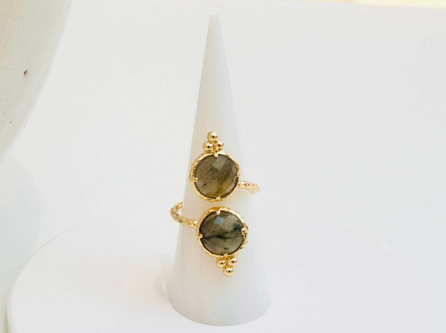 
                  
                    Two gold rings with oval labradorite stones displayed on a white cone ring holder over a round white base, with a large textured ceramic vessel partially visible on the left.
                  
                