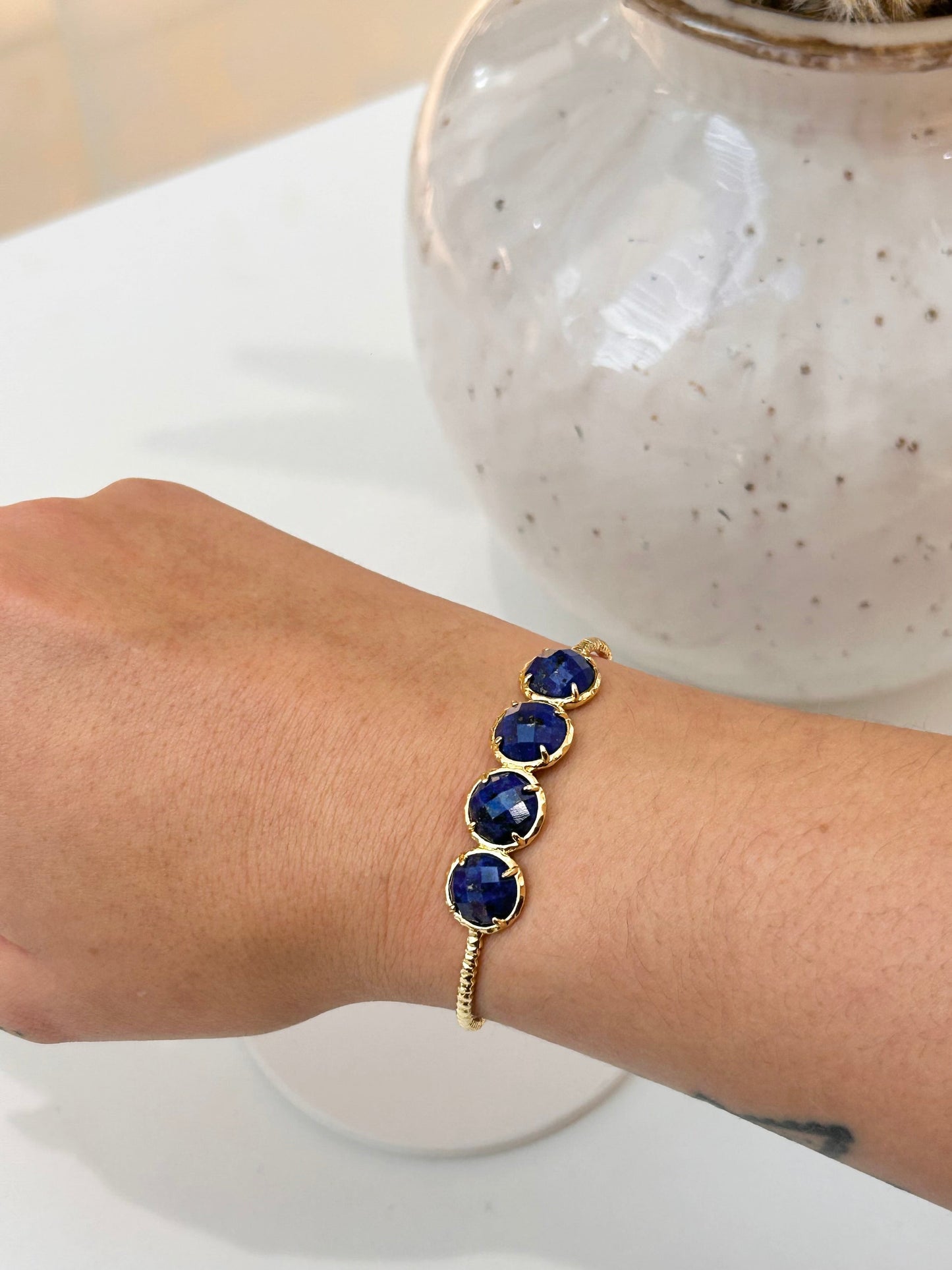 A gold bracelet with four round Lapis Lazuli gemstones worn on a person's wrist, shown against a white tabletop with a speckled ceramic vase in the background.