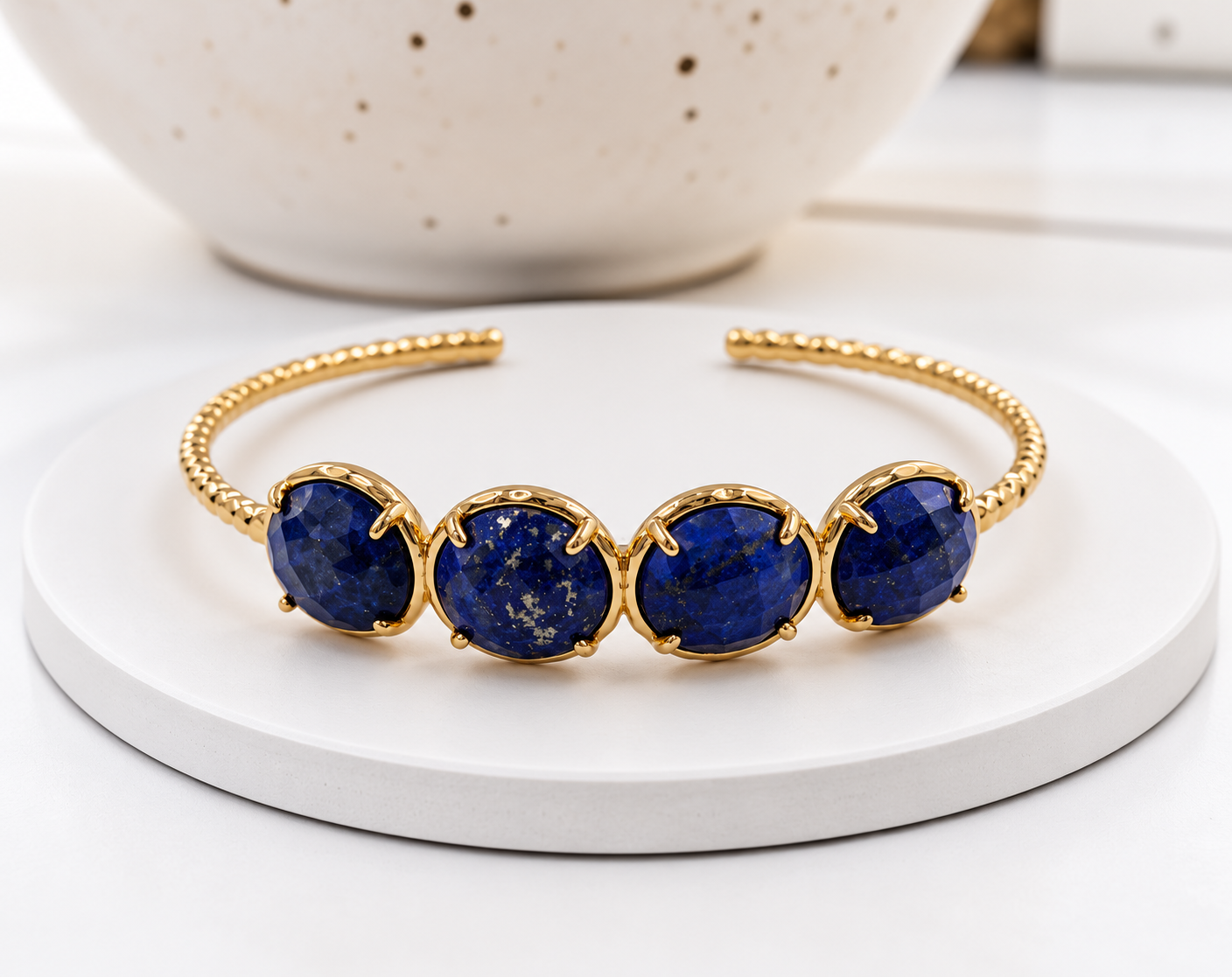 A gold cuff bracelet featuring four oval deep blue lapis lazuli stones, displayed on a white round pedestal with a speckled ceramic bowl in the background.