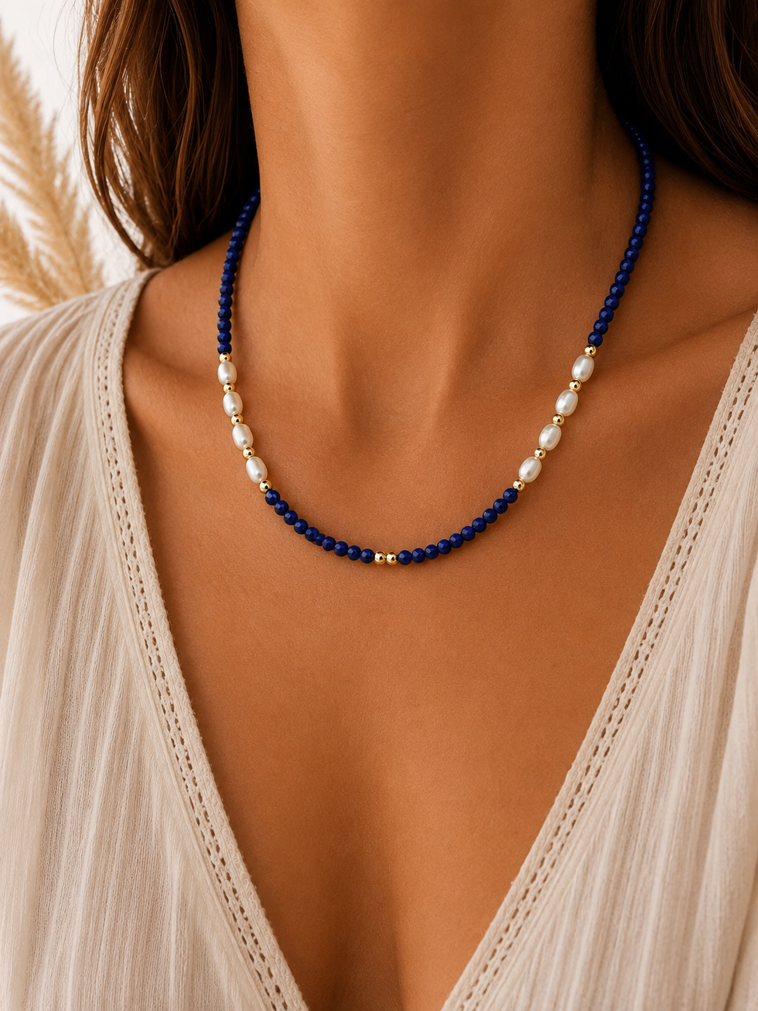 
                  
                    A close-up of a woman wearing a lapis lazuli beaded necklace with deep blue beads, white freshwater pearls, and small gold spacer beads, paired with a cream-colored lace-trim top.
                  
                