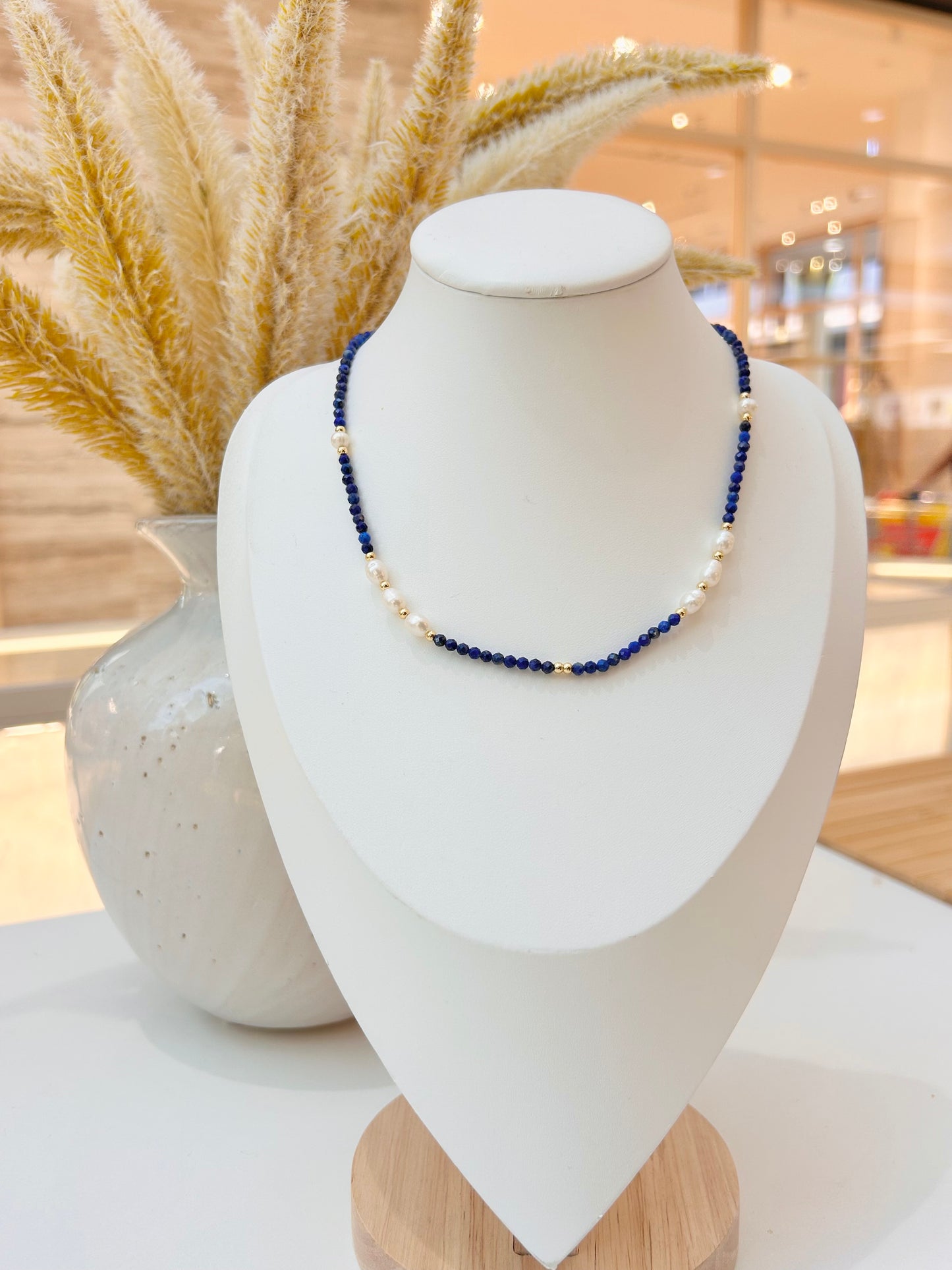 
                  
                    A beaded necklace with  blue lapis lazuli  and freshwater pearl beads displayed on a white jewellery bust, with dried pampas grass in a vase in the background.
                  
                