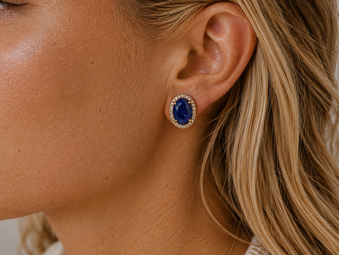 A close-up side profile of a woman wearing a gold oval stud earring with a deep blue lapis lazuli gemstone surrounded by a halo of small clear stones.