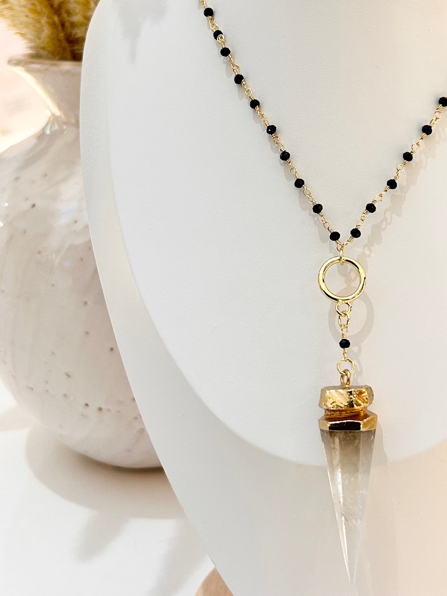 
                  
                    A beaded necklace displayed on a white mannequin bust, featuring black and clear faceted beads with a gold-toned ring and a clear pointed crystal pendant.
                  
                