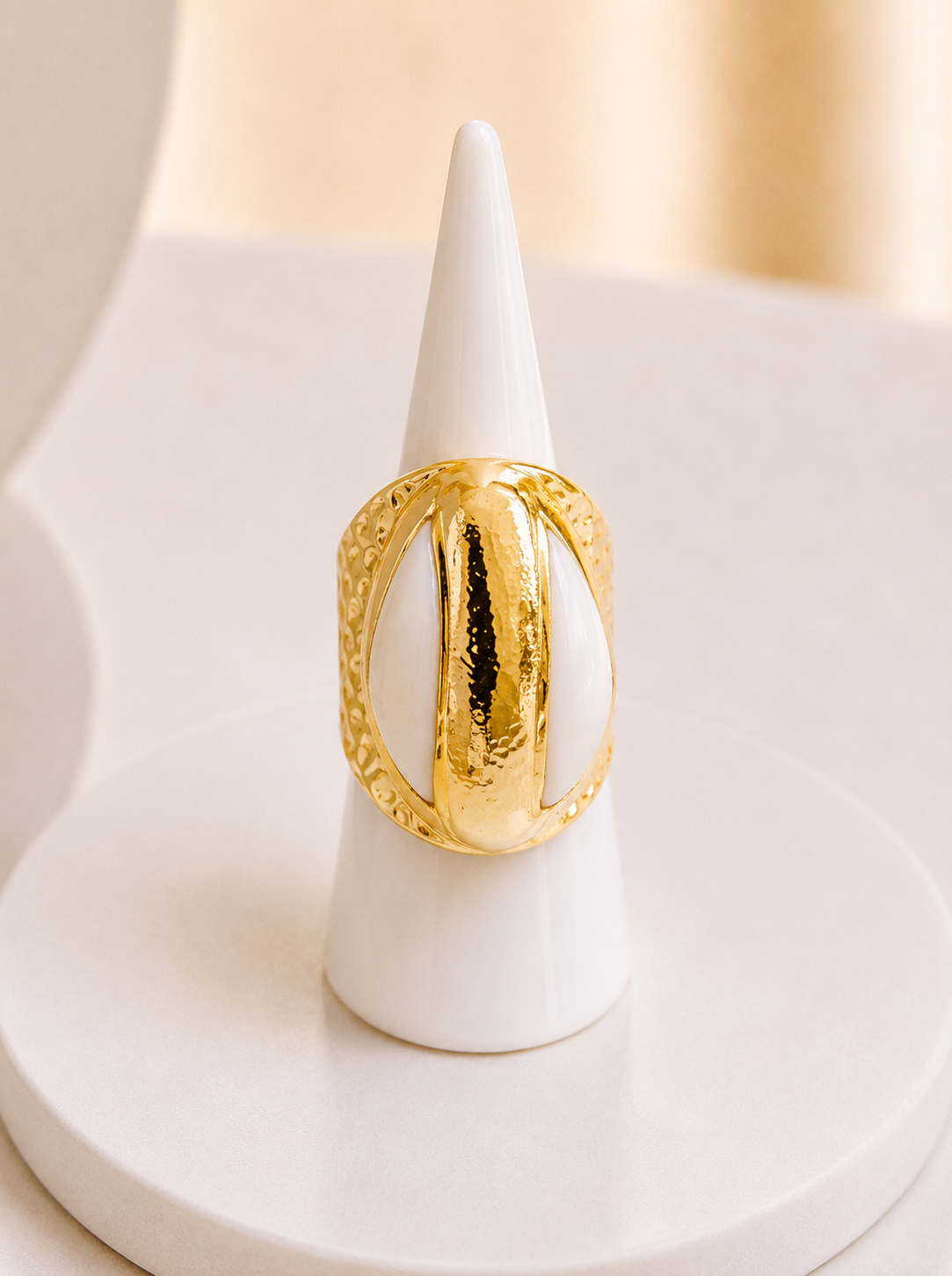 A chunky gold ring with a textured hammered finish and a central polished gold bar, displayed on a white cone jewelry stand.