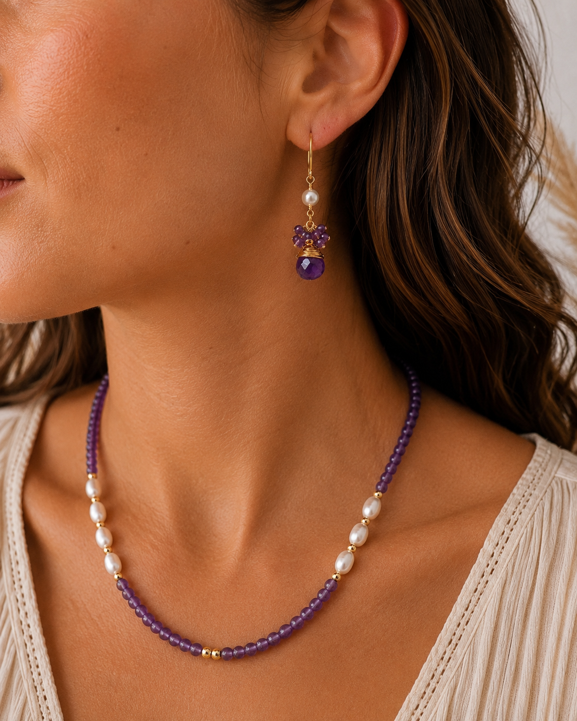 
                  
                    A close-up lifestyle shot of a woman wearing a matching purple amethyst beaded necklace and drop earring. The jewelry features purple amethyst beads, white freshwater pearls, and gold-toned metal accents.
                  
                