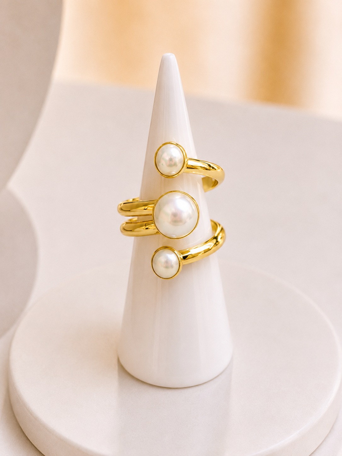 Three gold freshwater pearl stones on an adjustable ring, displayed on a white cone jewelry stand against a warm beige background.