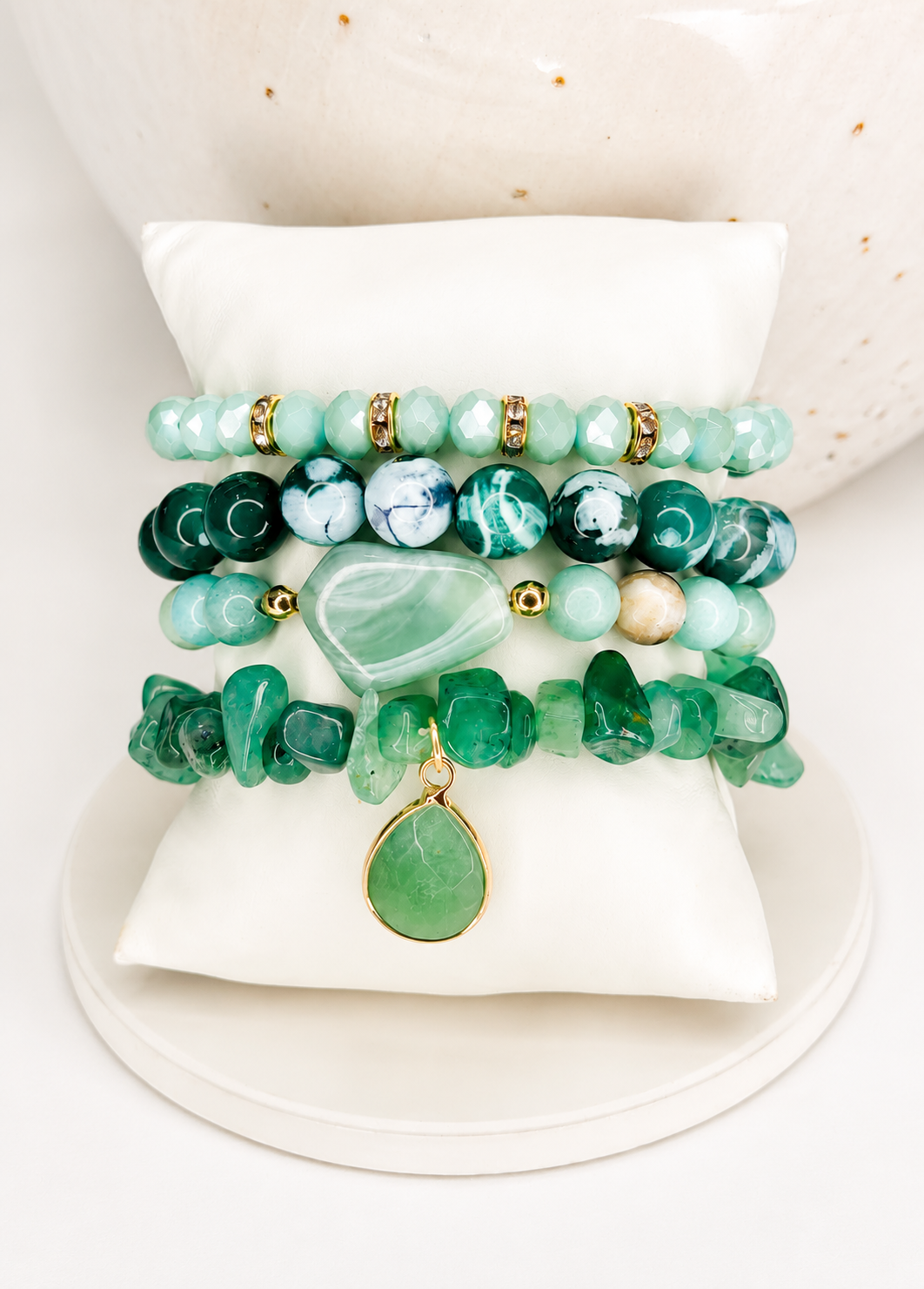 Four stacked beaded and polished stone bracelets in shades of green, teal, and white, with gold spacer natural beads and a green teardrop pendant, displayed on a white cushion.