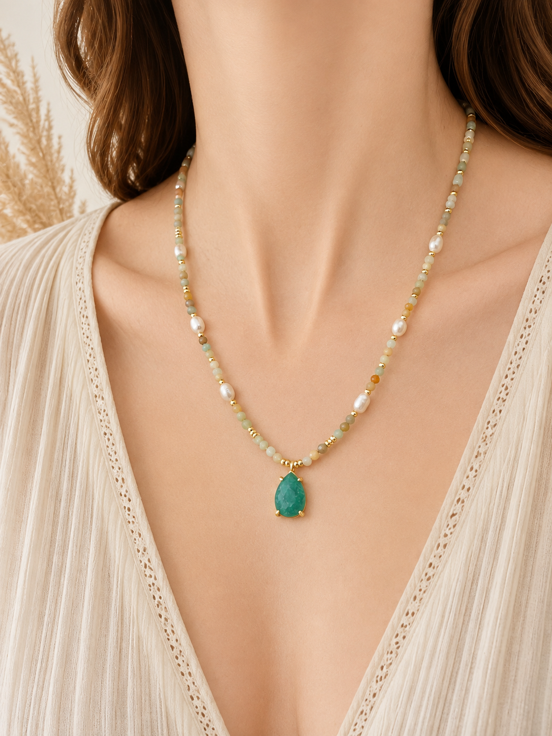 A close-up of a woman wearing a delicate beaded necklace with a amazonite teardrop pendant. The necklace features small white pearls, light green beads, and gold-toned details, styled with a cream-colored lace-trim top.