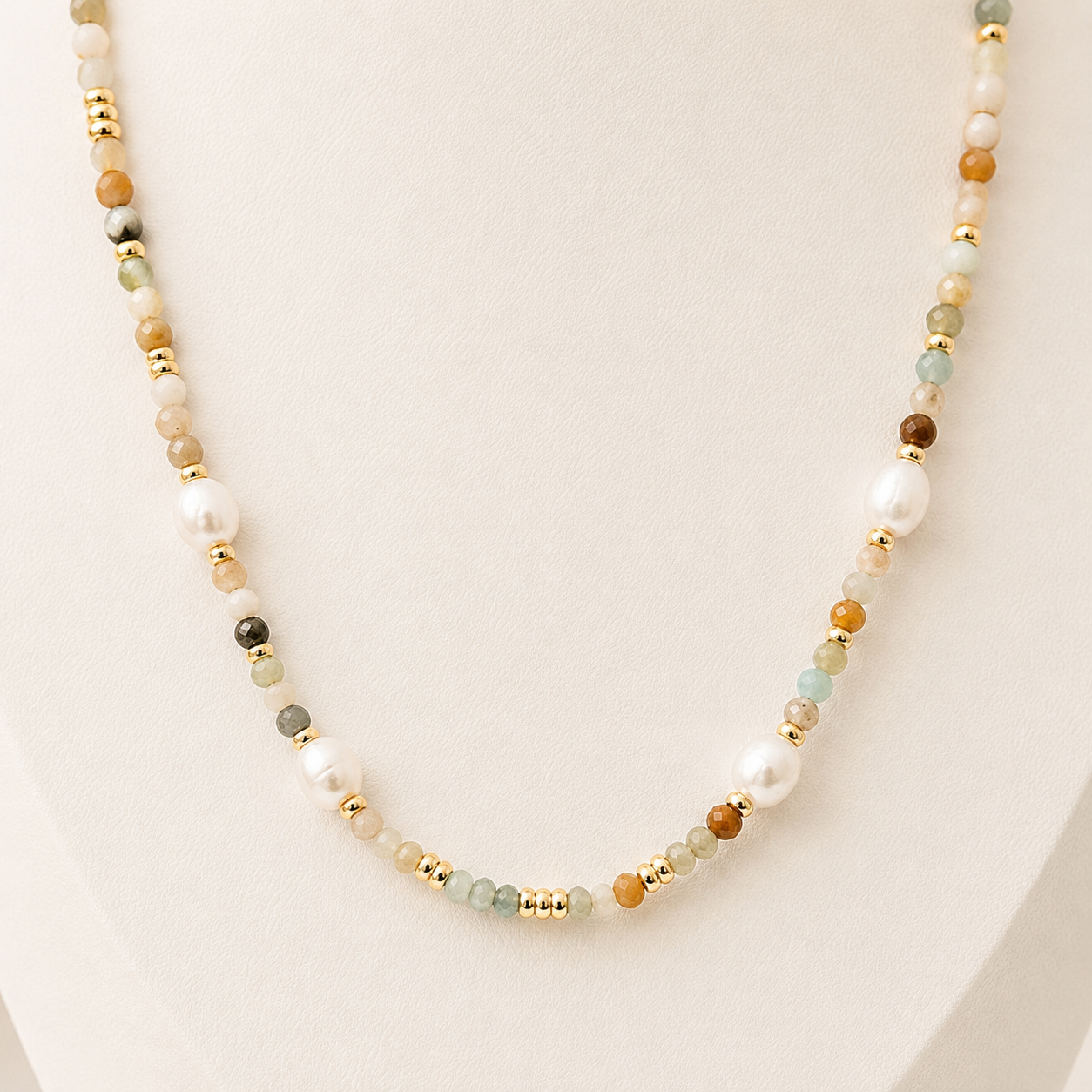 A beaded necklace displayed on a cream-colored jewellery bust. The necklace features a mix of white freshwater pearls, small gold spacer beads, and various mixed amazonite stone beads in brown, light green, and grey.