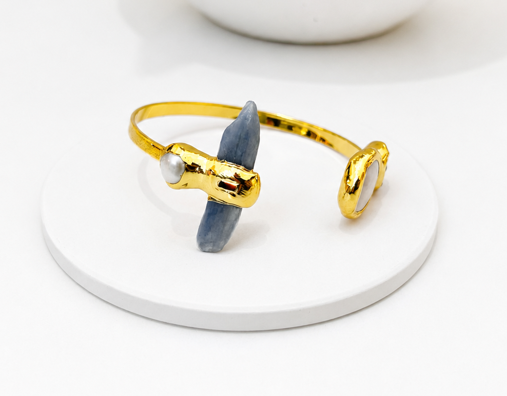 
                  
                    A gold-tone open cuff bracelet with a blue-gray stone and a small white pearl accent, displayed on a round white coaster.
                  
                