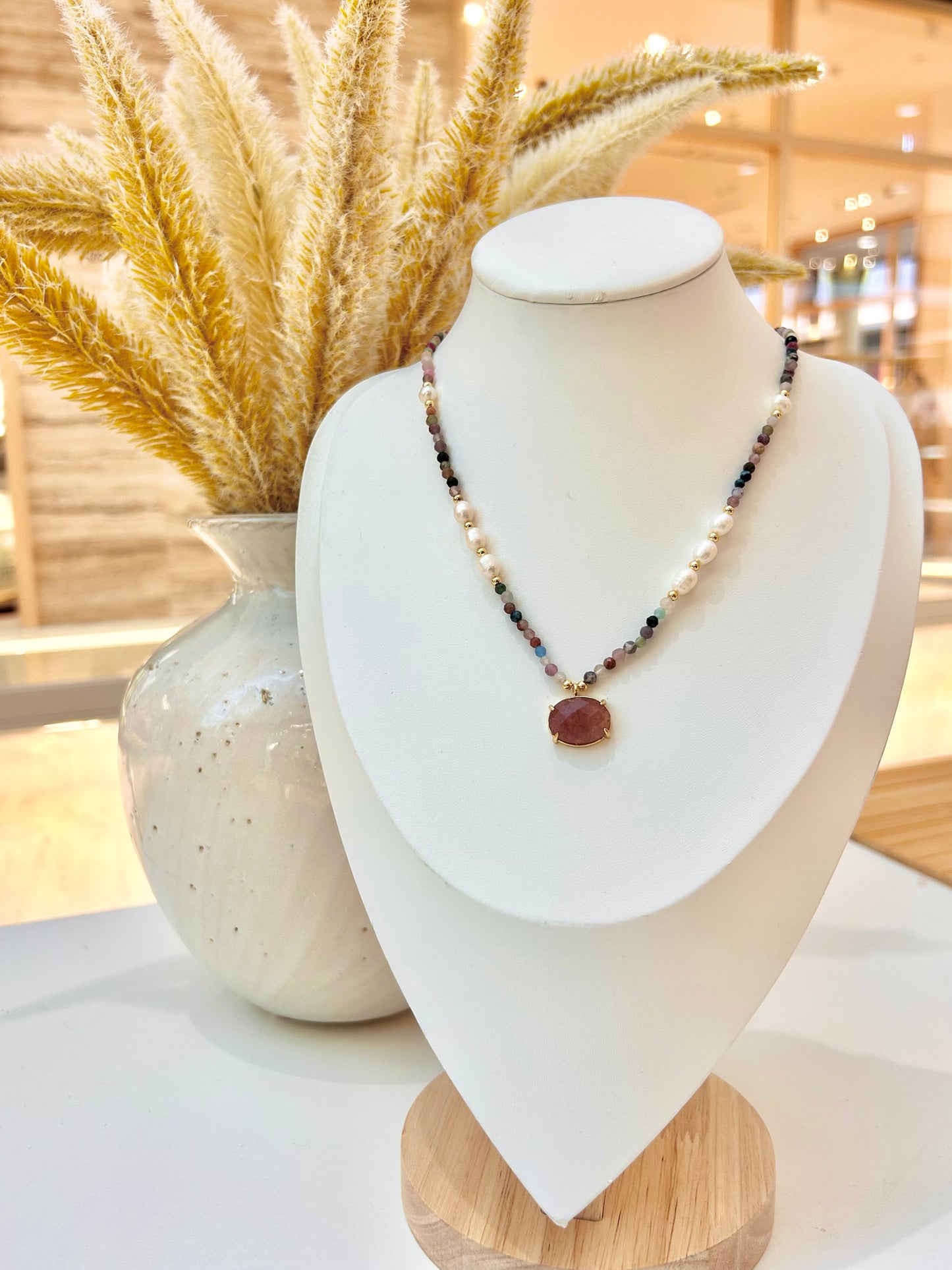 
                  
                    A beaded necklace with a strawberry quartz oval pendant displayed on a white jewellery bust. The necklace features white, dark blue, and metallic beads, styled in front of a ceramic vase with dried pampas grass in a softly blurred indoor setting.
                  
                