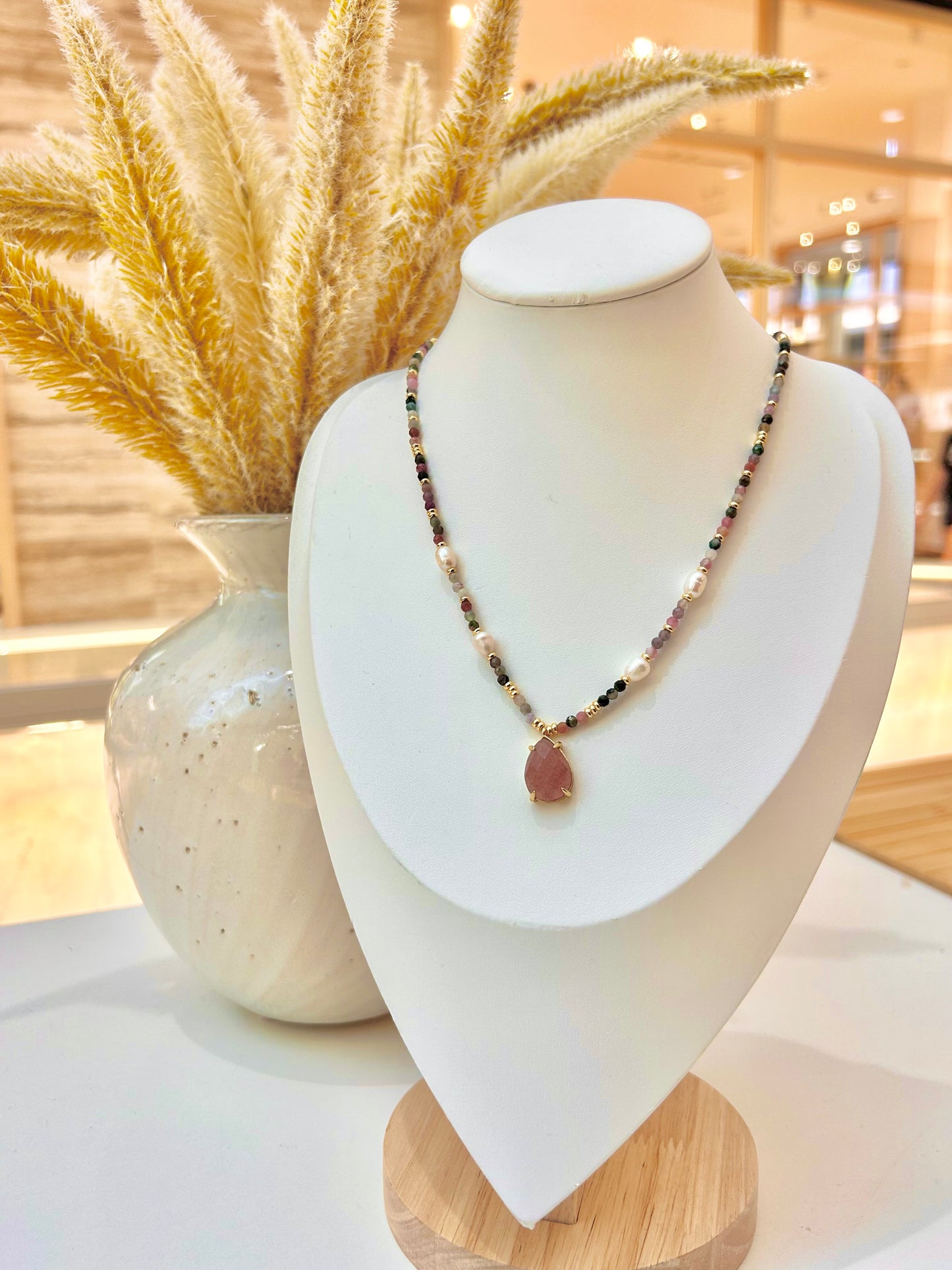 
                  
                    A beaded necklace with a teardrop-shaped sunstone pendant displayed on a white jewellery bust. The necklace features small dark and light beads with a gold-toned setting.
                  
                