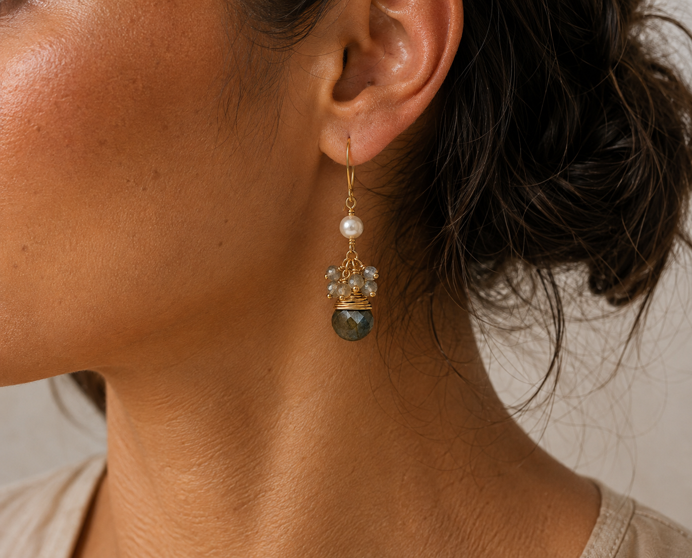 A close-up of a model's ear wearing a gold dangle earring with a white pearl, clear crystals, and a faceted Labradroite stone.