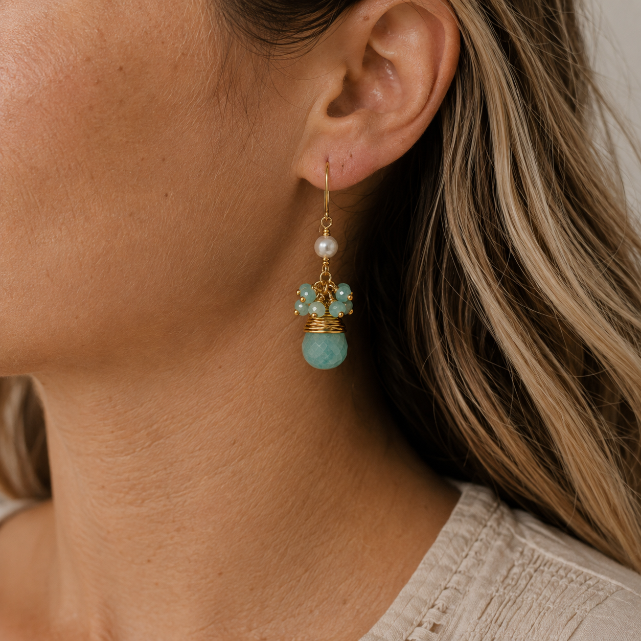 A close-up of a woman's ear wearing a dangling earring. The earring features a gold hook, a small white pearl, several small amazonite beads, and a larger polished Amazonite blue bead at the bottom.