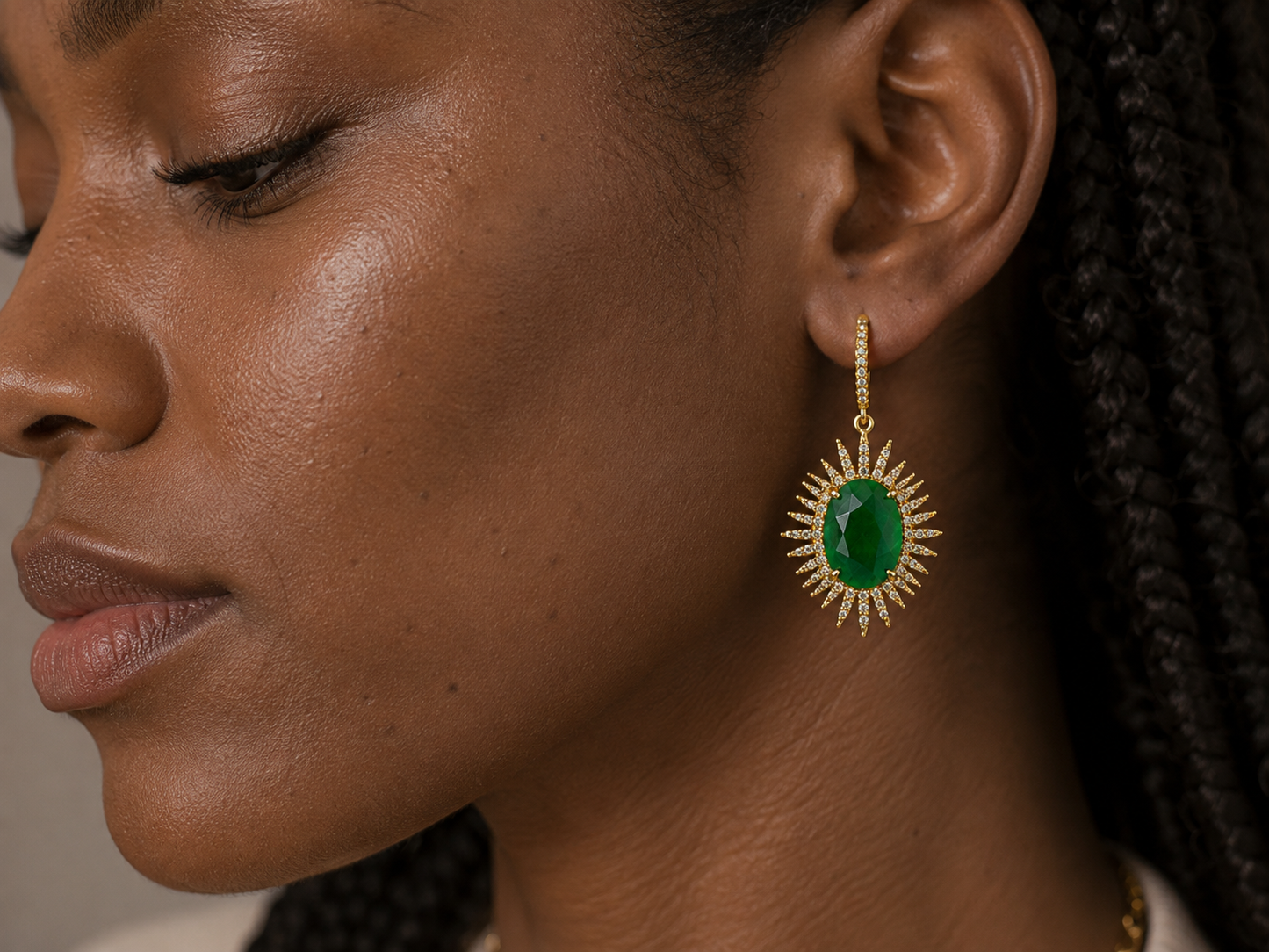 A close-up side profile of a woman wearing a gold sunburst dangle earring with a green oval center stone.
