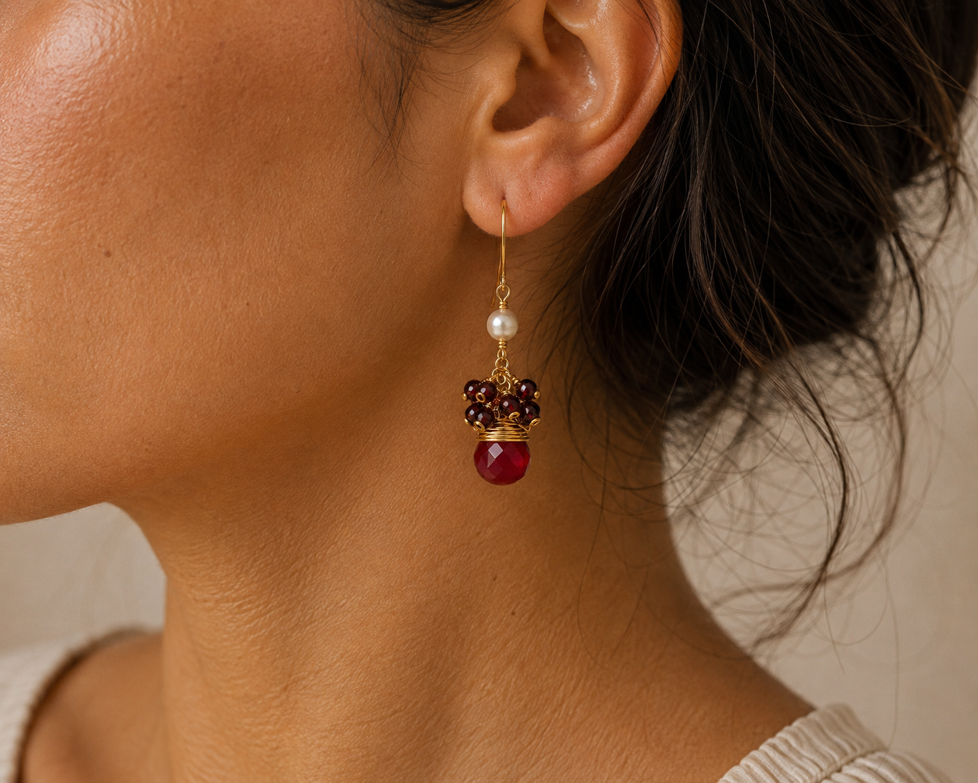 A close-up of a model's ear wearing a gold dangle earring with a white pearl, dark brown beads, and a deep red jade bead.