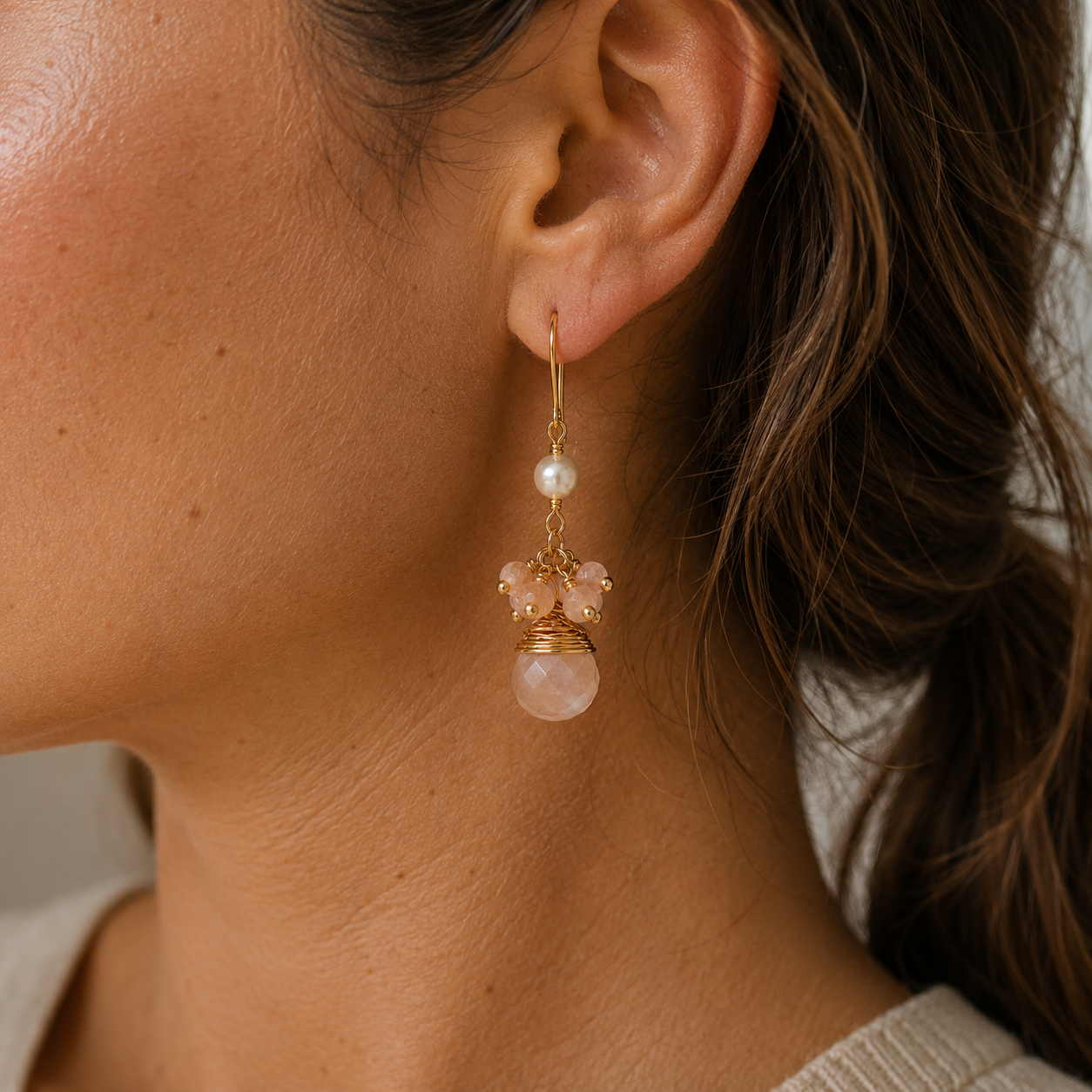 A close-up of a model's ear wearing a delicate gold dangle earring with a white pearl, pink sunstone beads, and a large round pink sunstone.