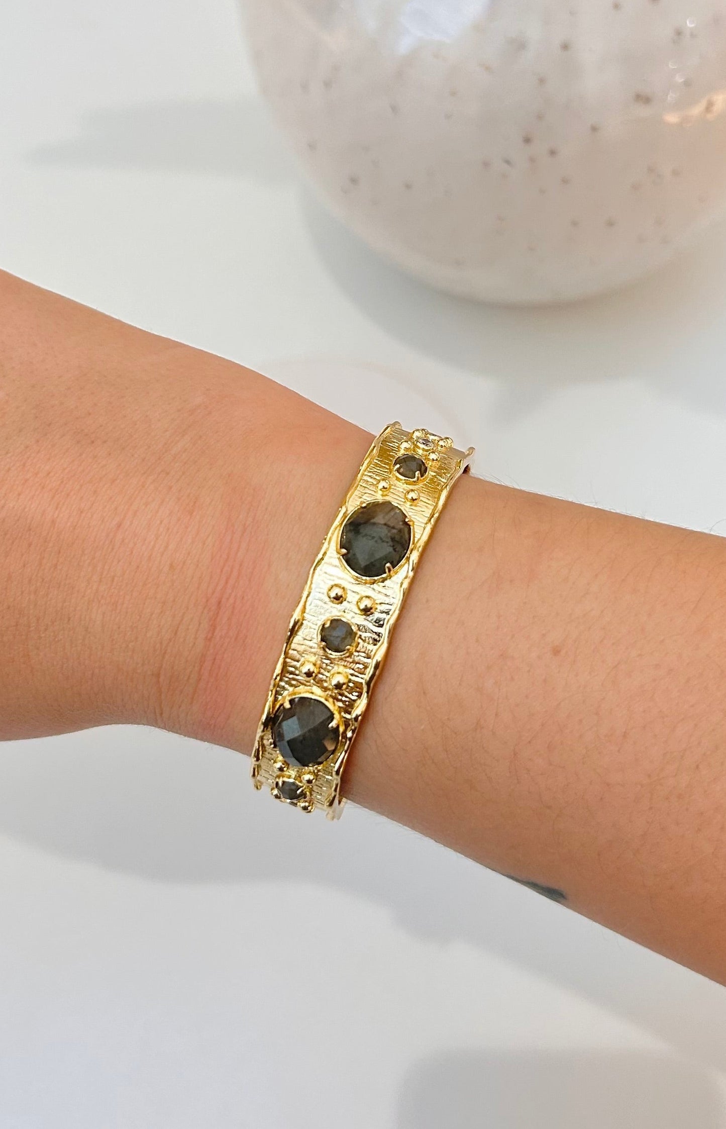 A gold-toned cuff bracelet with dark oval stones, worn on a person's wrist, with a ceramic vase in the background.