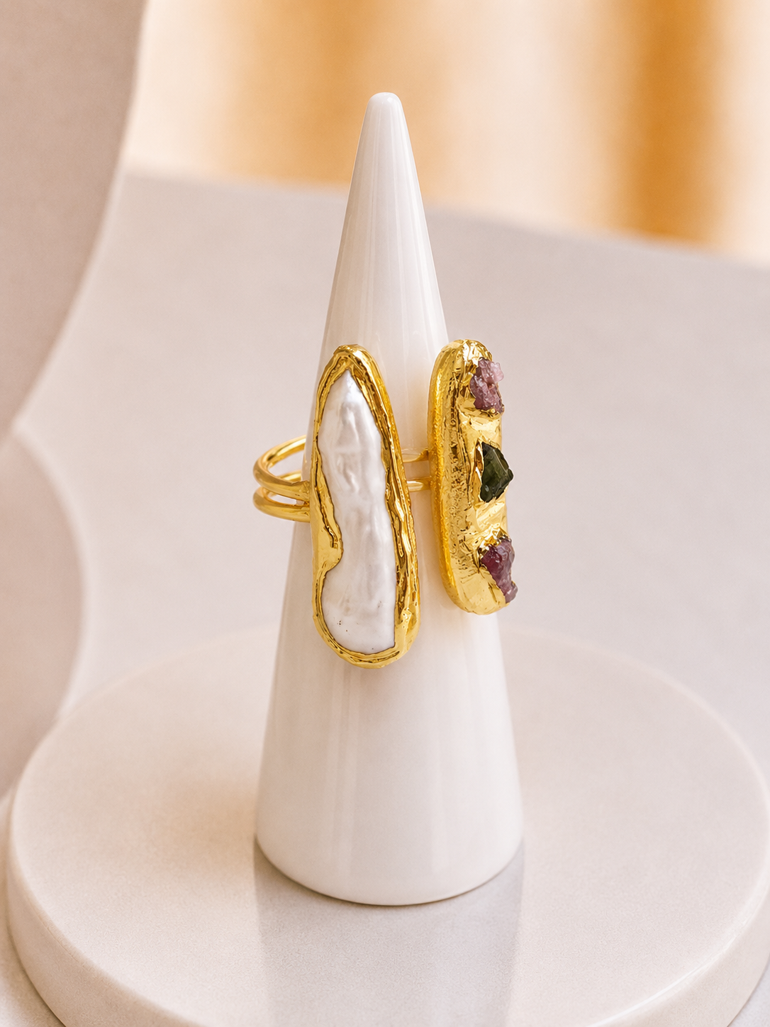 A gold ring displayed on a white cone stand, featuring a large white textured stone on one side and a smaller side panel with purple and green stones on a gold base.